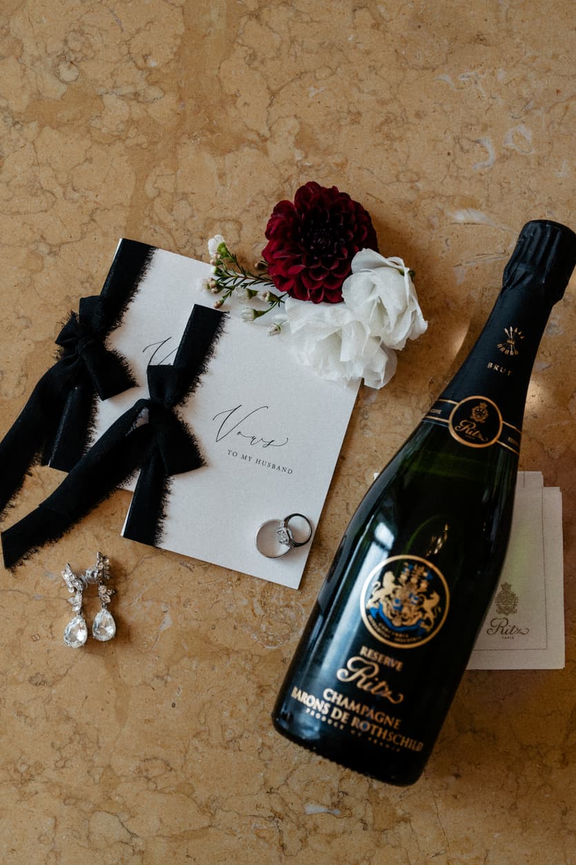 Wedding details on a marble table: vow book, rings, earrings, champagne bottle, ribbons, and white flowers