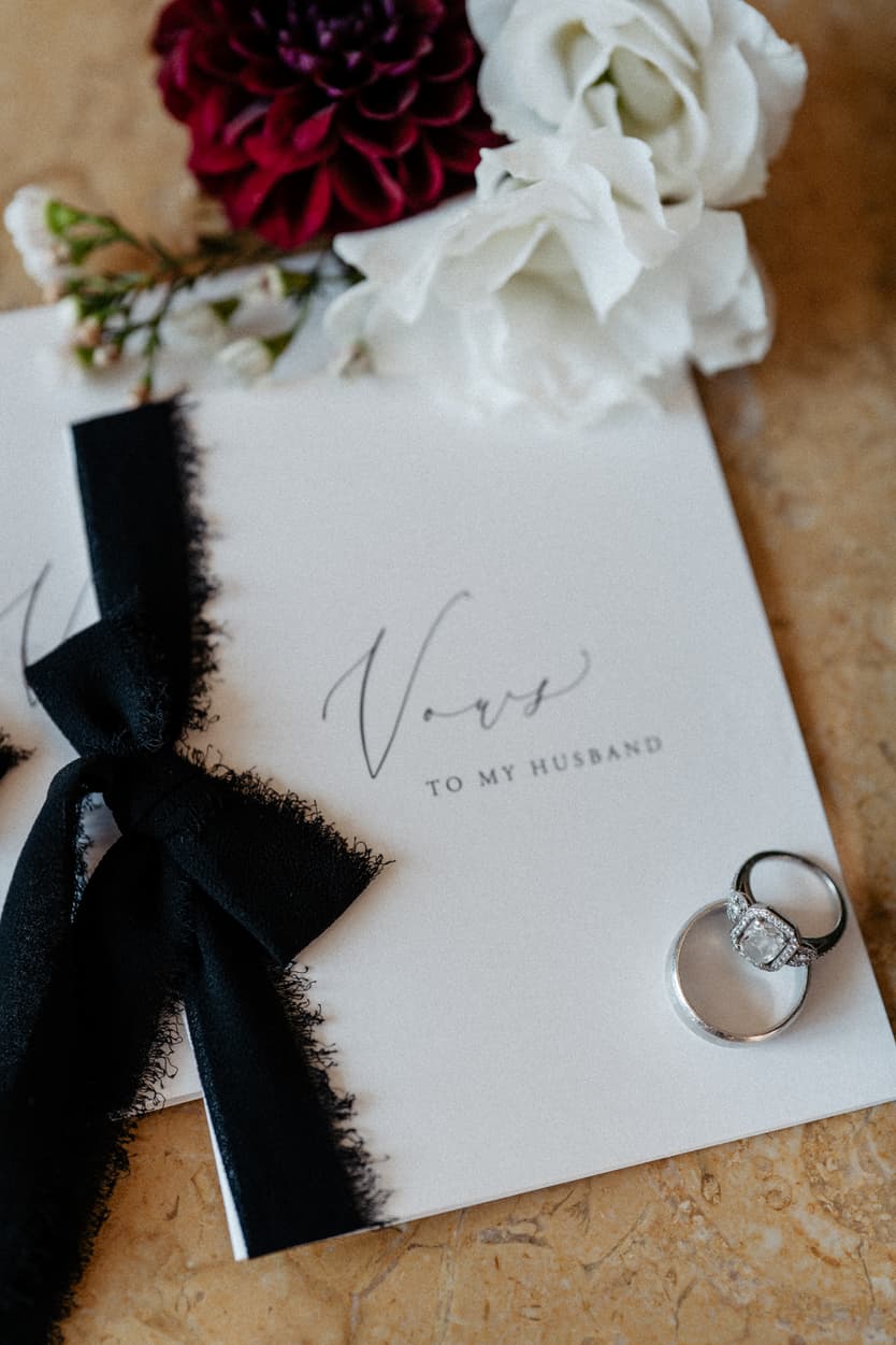 Wedding vow book with black ribbon, rings, and white flowers on a warm tabletop in soft light