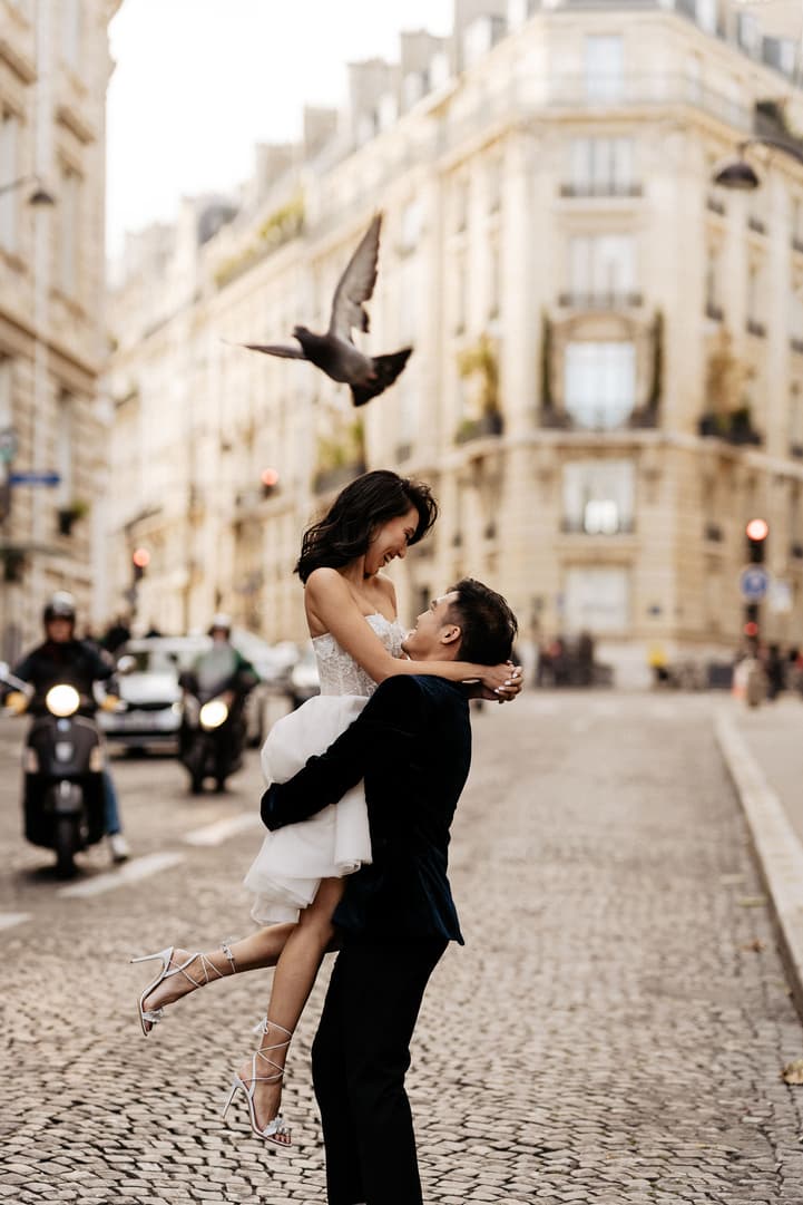 Bride in white dress lifts with groom on a Paris street at golden light, scooters passing, a bird overhead