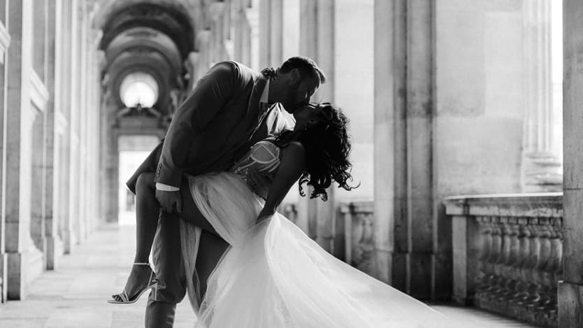 Bride and groom kiss in a grand arched colonnade, her veil flowing in soft light