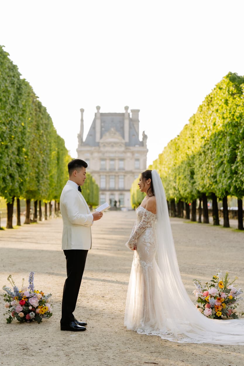 Bride and groom face each other in warm light in a tree-lined garden, with vows in hand and flowers at their feet