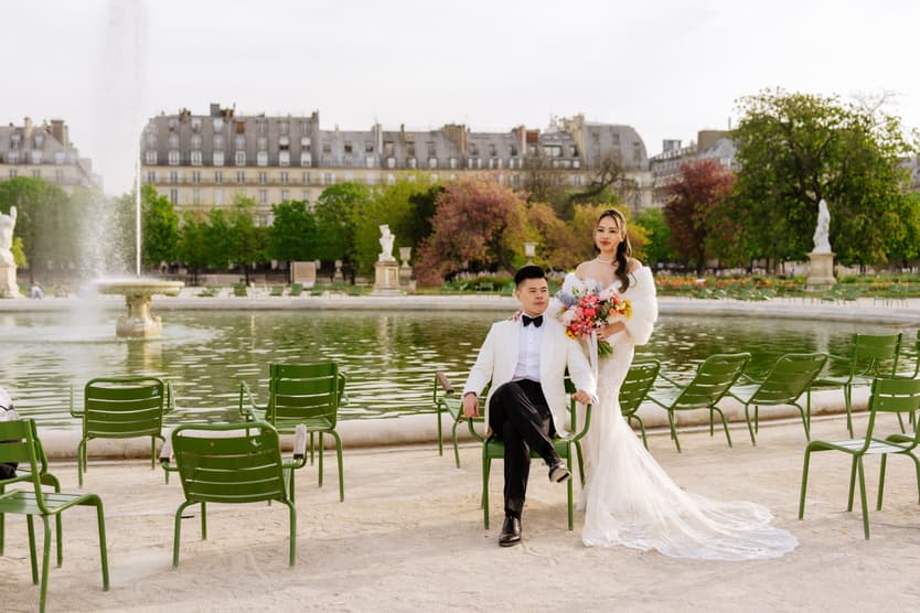 Bride and groom pose by a fountain and pond in a Paris garden, with green chairs and soft daylight