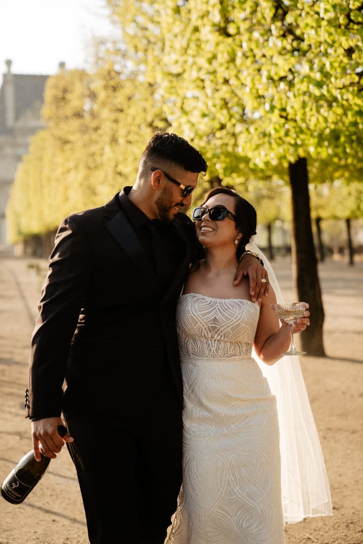 Bride and groom stroll through a sunlit Paris garden, arm in arm, holding champagne bottle and coupe glasses