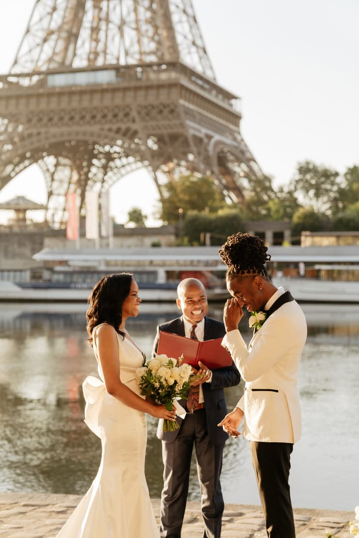 Bride and groom with an officiant by the Seine, a red vow folder open beneath the Eiffel Tower