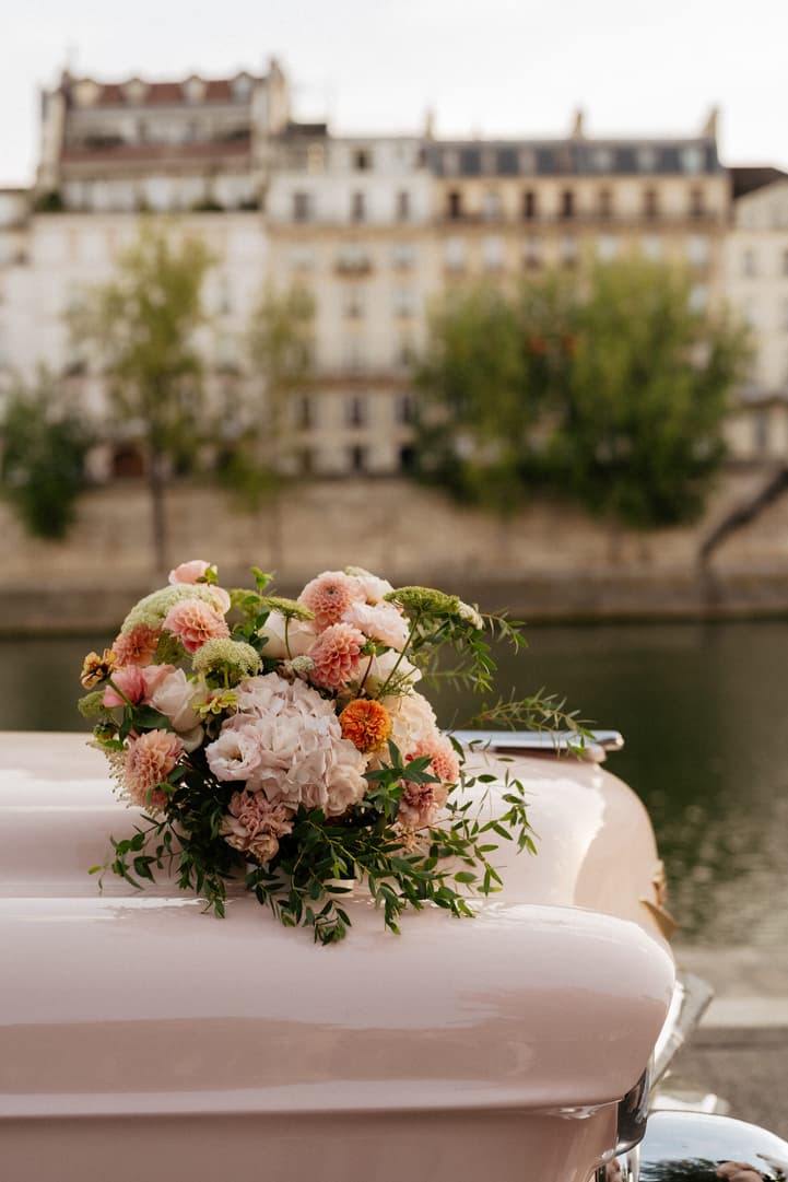 A pastel bridal bouquet rests on a pink car by the Seine, with Paris buildings softly blurred behind