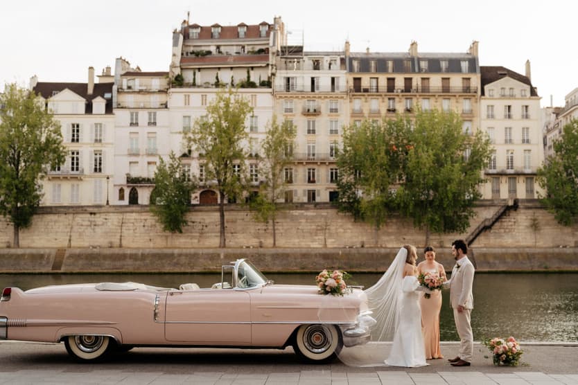 Bride and groom stand beside a pink vintage car on the Seine, with soft light and Paris apartments behind