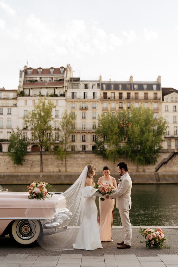 Bride and groom exchange bouquets with an officiant beside a pink car on the Seine under soft light