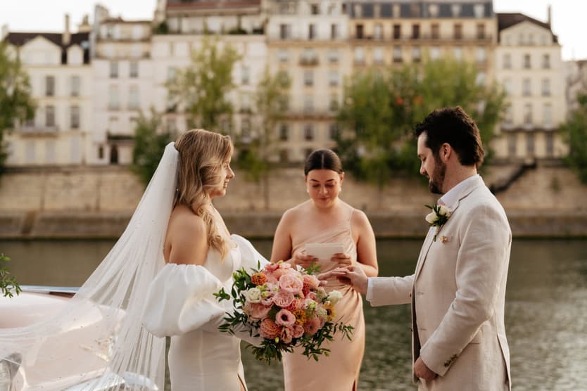Bride and groom exchange rings beside the Seine with an officiant behind them and a bouquet in warm light