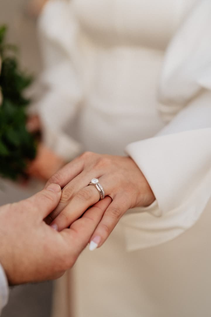 Bride and groom clasp hands during a wedding ceremony, showing a diamond ring and white sleeves