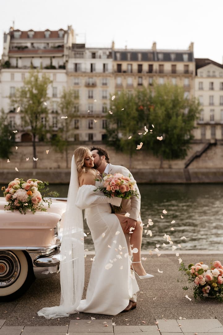 Bride and groom kiss on a Paris riverbank beside a pink vintage car, with petals drifting in soft light