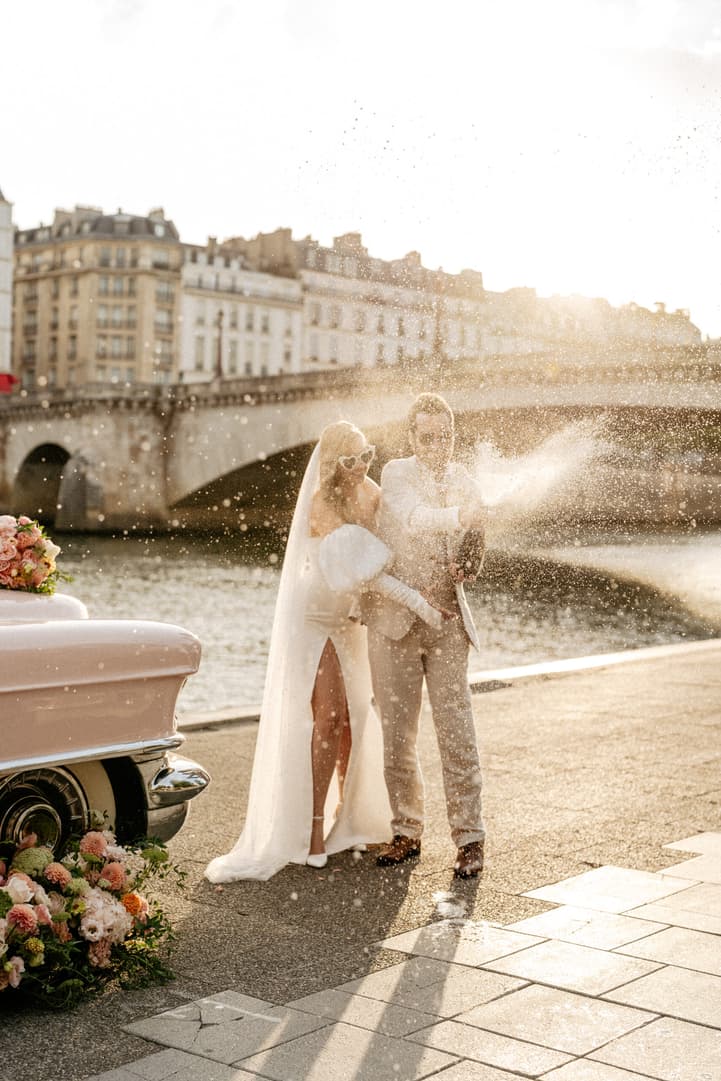 Bride and groom on the Seine in warm light, spraying champagne beside a flowered vintage car