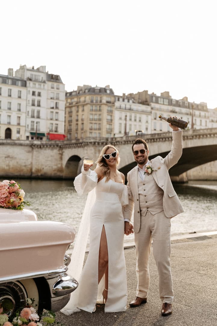 Bride and groom toast by the Seine beside a pink vintage car in warm sunset light