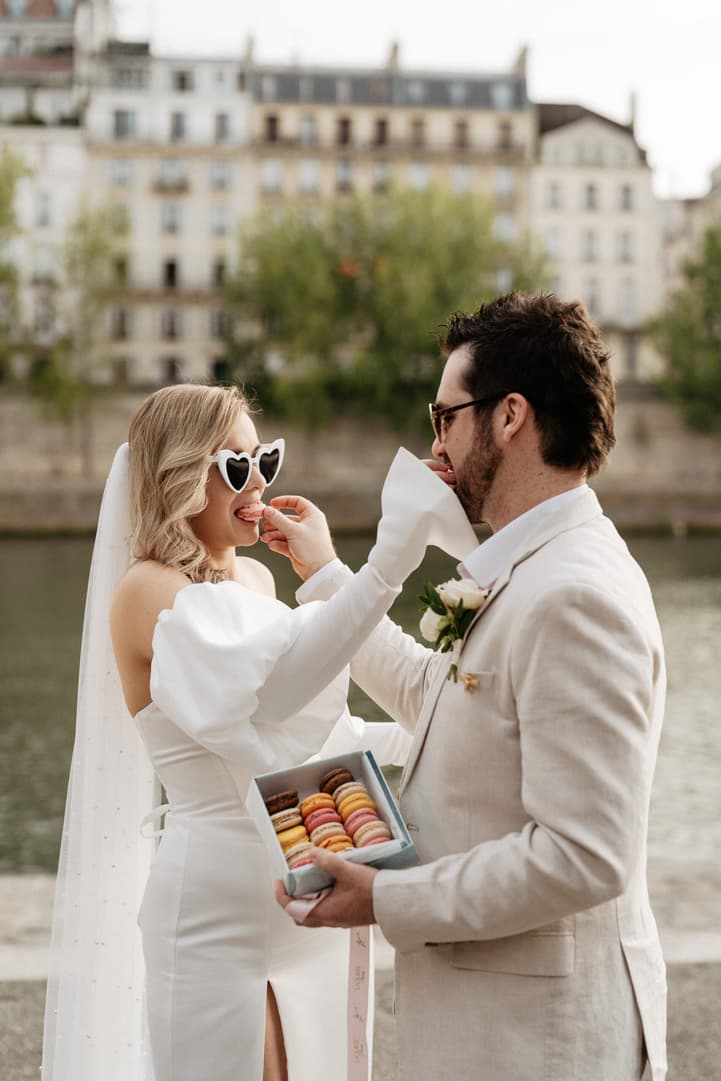 Bride and groom share a macaron on a Paris riverbank, with soft light and a box of colorful macarons