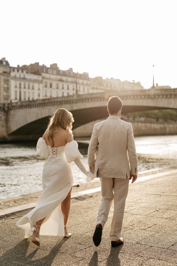 Bride and groom walk hand in hand along the Seine at sunset, with a stone bridge behind them