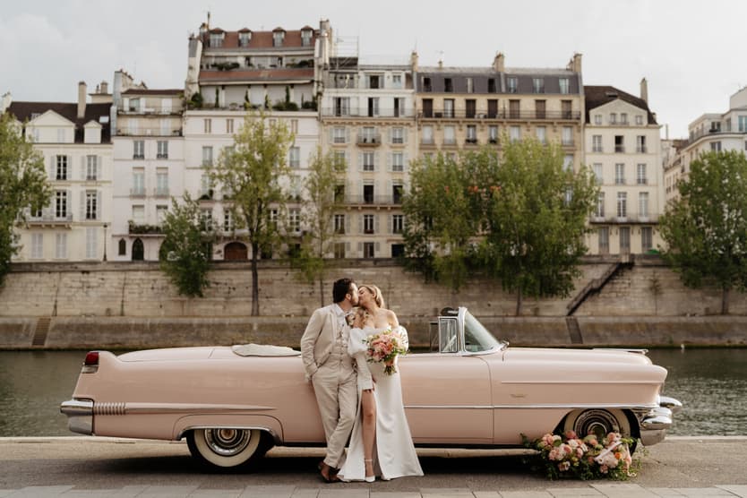 Bride and groom pose beside a pink vintage convertible on the Seine, with bouquets and Paris flats behind