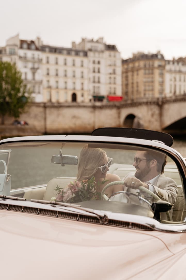 Bride and groom smile in a vintage convertible on a Paris street by the river in soft light