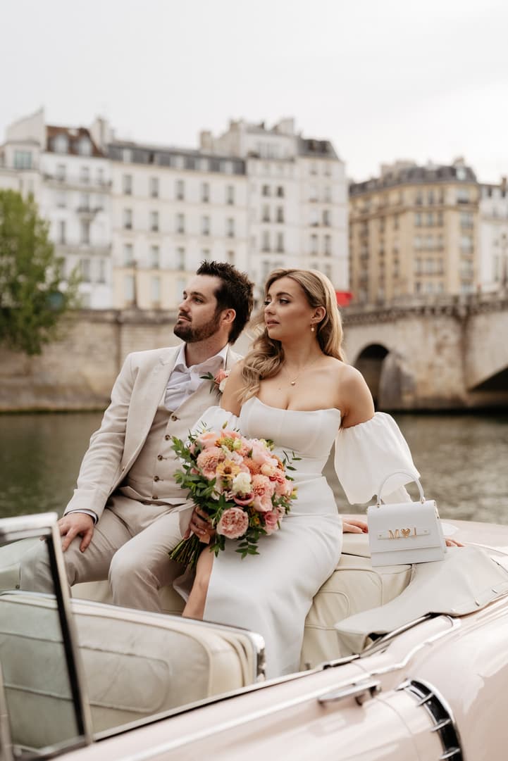 Bride and groom sit in a boat on the Seine, holding a bouquet in soft daylight with Paris buildings behind