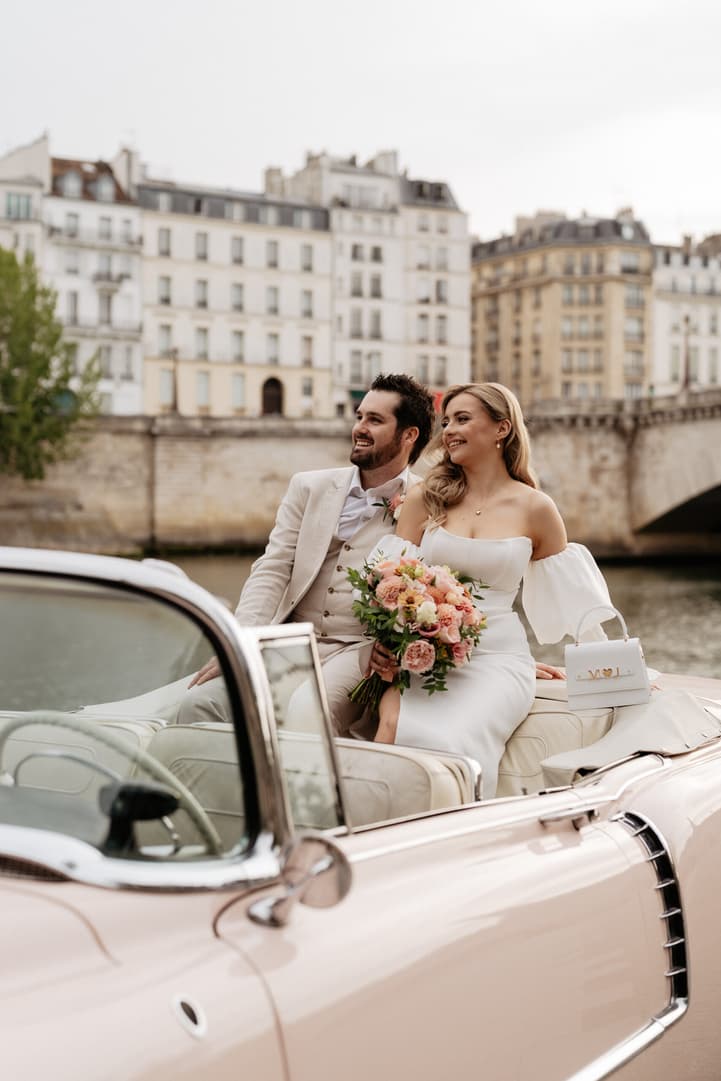 Bride and groom sit in a vintage convertible by the Seine, with a pastel bouquet and stone bridge behind