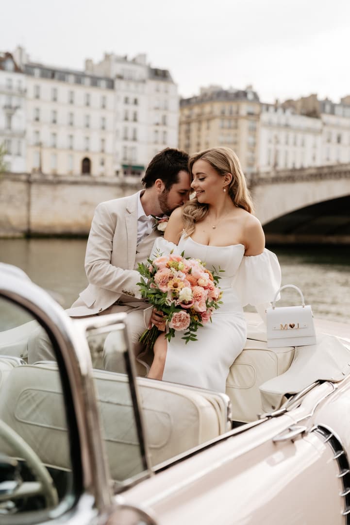 Bride and groom share a quiet moment in a vintage car by the Seine, with pastel Paris buildings behind