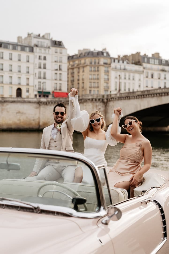 Bride, groom and guest raise hands in a pink convertible by the Seine in soft light, wearing heart sunglasses