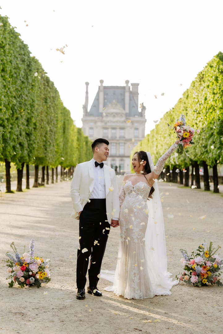 Bride and groom hold hands in a sunlit Paris garden as petals fall before a stately palace