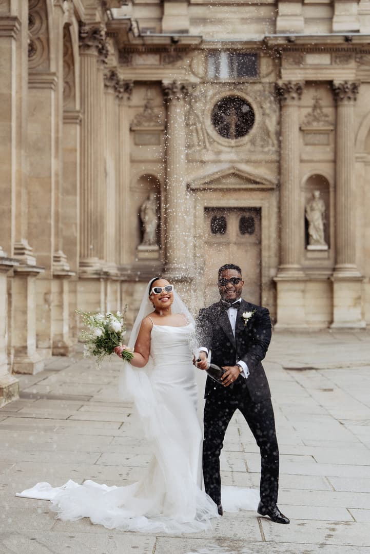 Bride and groom spray champagne in a sunlit stone courtyard outside a church