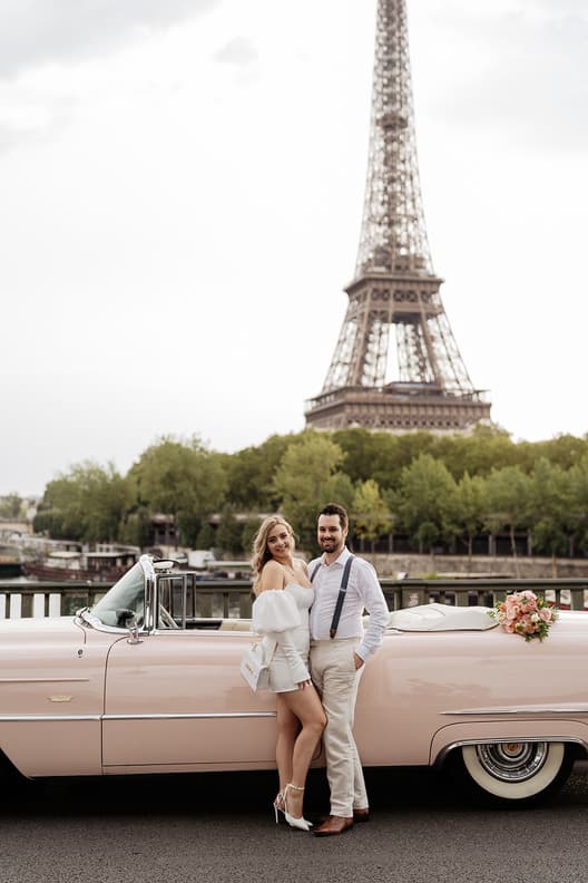 Bride and groom pose by a pink vintage car with a bouquet, the Eiffel Tower rising behind them in soft light