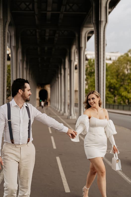 Bride and groom hold hands on a Paris street under a bridge in soft light, with her white dress and bag