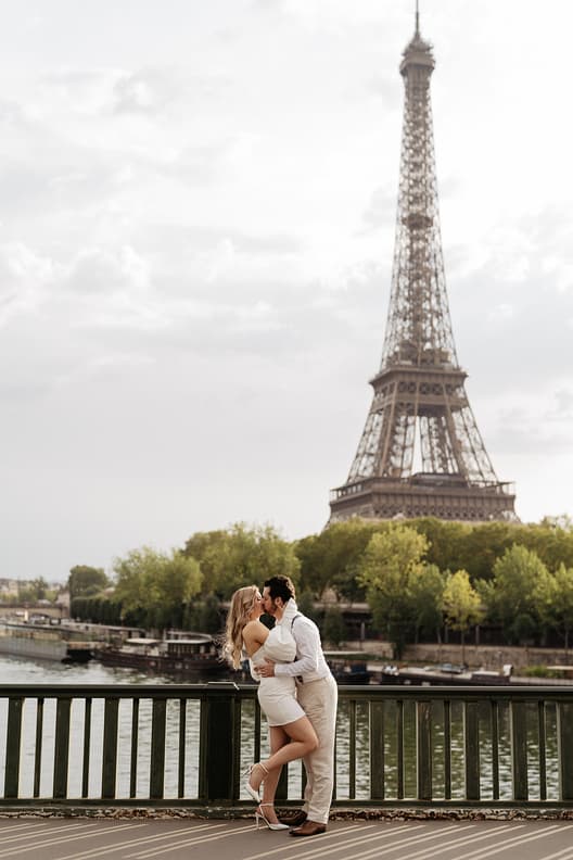 A couple kisses by the Seine with the Eiffel Tower rising above the trees in soft daylight