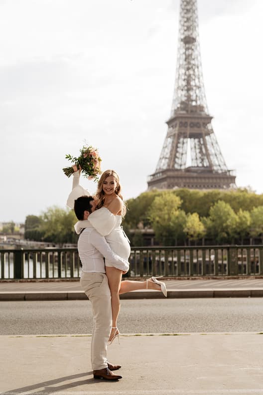 Bride lifts bouquet as groom hugs her by the Eiffel Tower in soft daylight on a Paris street