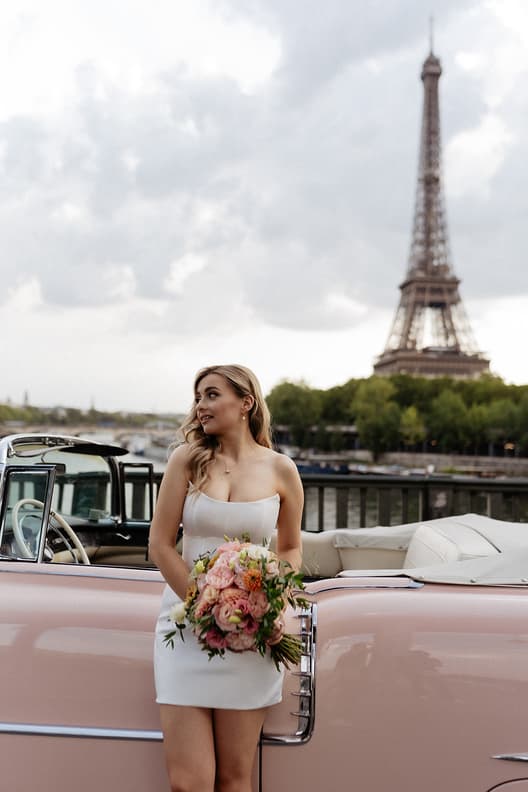 Bride in a white dress holds a pink bouquet beside a vintage car on the Seine with the Eiffel Tower behind