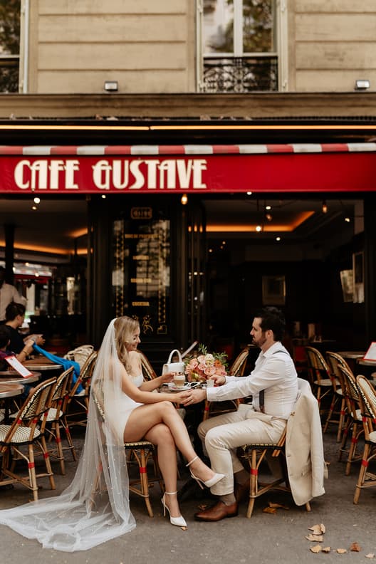 Bride and groom sit hand in hand at a Paris street café, with a bouquet and warm evening light
