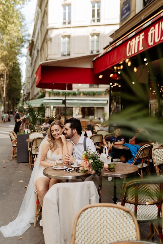 Bride and groom sit at a Paris street café, sharing coffee in soft daylight beside red awnings