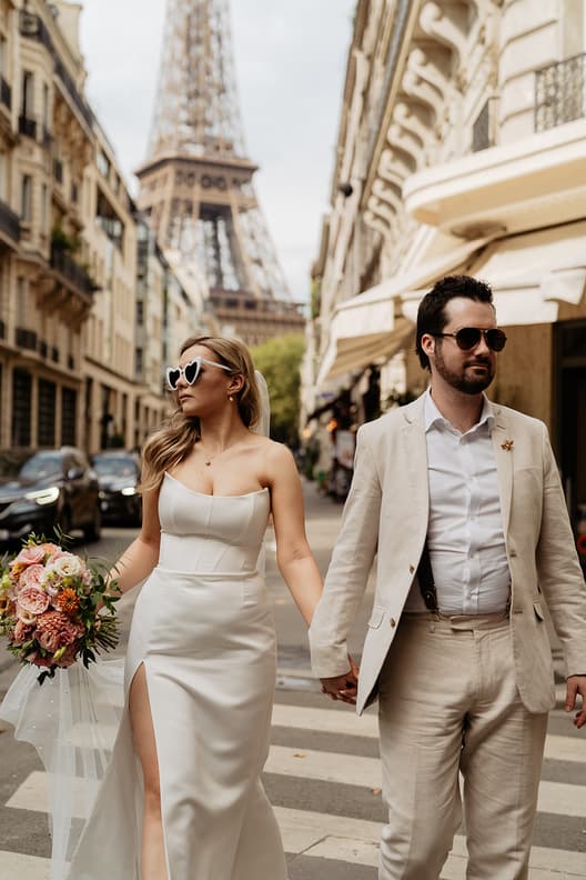 Bride and groom hold hands on a Paris street with the Eiffel Tower behind them and a bouquet at her side
