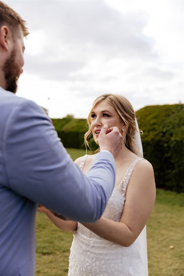 Groom gently adjusts the bride’s face in a green garden under soft cloudy light