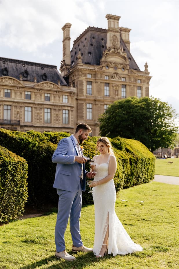 Bride and groom pour champagne in a sunlit garden before the Louvre facade
