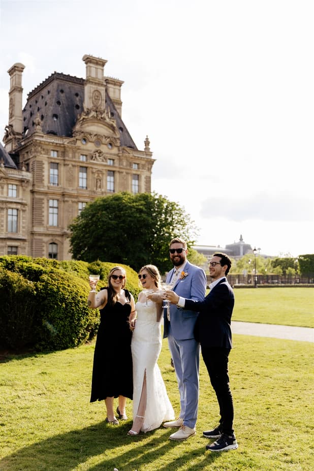 A bride and groom toast with two guests in a Paris garden beside an ornate palace facade in bright sun