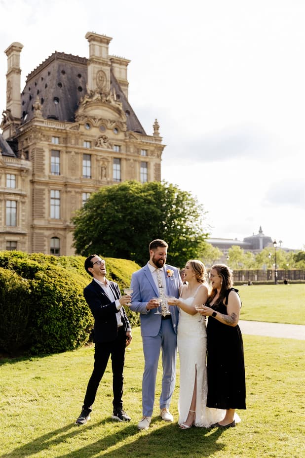 Bride and groom toast with two guests on a sunny Paris garden lawn beside a grand historic palace