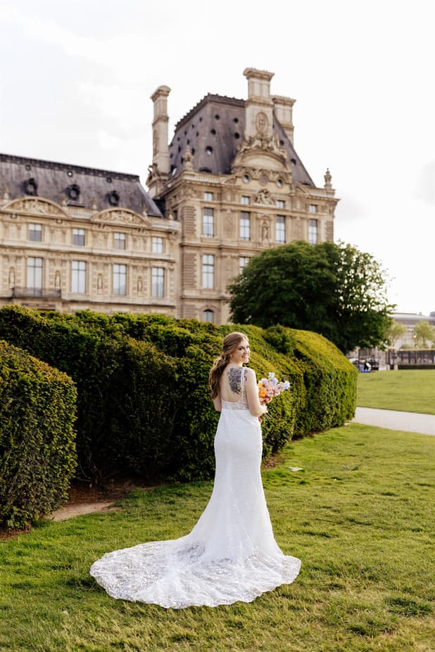 Bride in a lace gown holds a bouquet in a Paris garden before a grand museum, soft daylight above