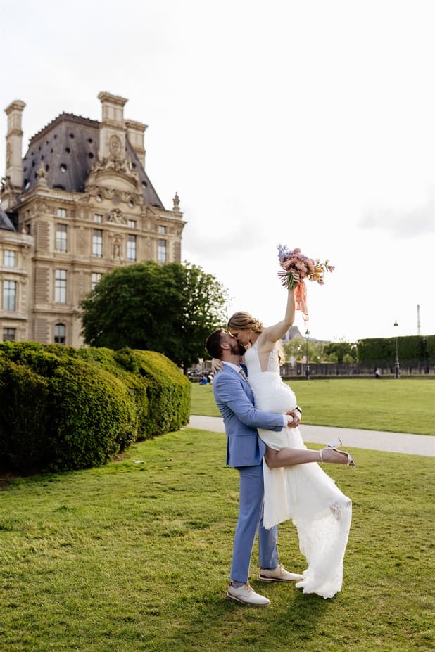 Bride and groom kiss in a Paris garden, her bouquet raised beside a grand historic palace