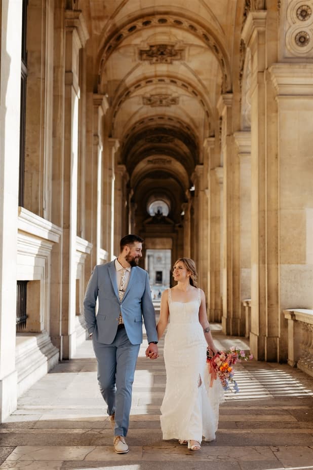 Bride and groom walk hand in hand through a sunlit Paris colonnade, bouquet in her other hand