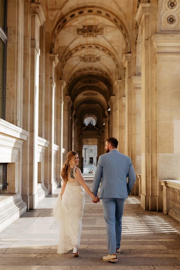 Bride and groom walk hand in hand through a grand stone arcade in warm Paris light