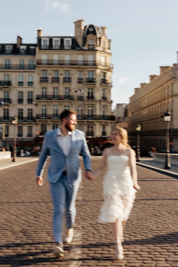 Bride and groom walk hand in hand on a Paris street in warm light, his blue suit and her ruffled dress swaying