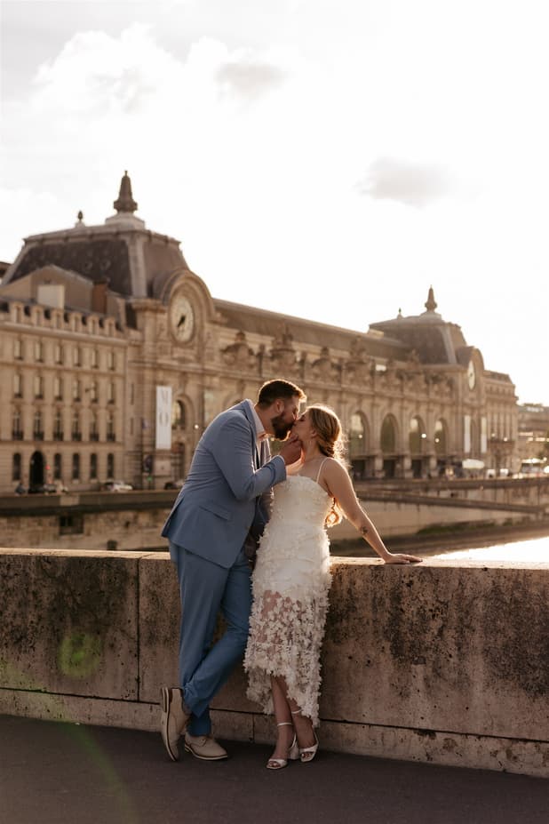 Bride and groom kiss by the Seine in warm backlight, with a historic station behind them