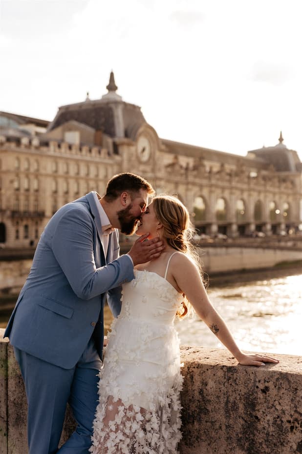 Bride and groom kiss by the Seine, backlit beside the Louvre's riverside façade