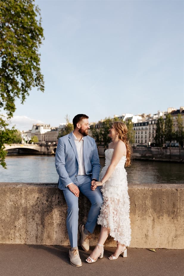 Bride and groom in blue suit and white dress sit by the Seine, holding hands in warm afternoon light
