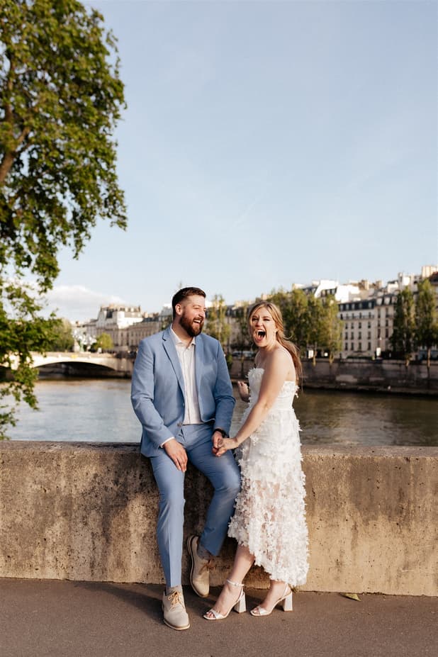 Bride and groom laugh by the Seine in soft evening light, hand in hand beside a stone quay