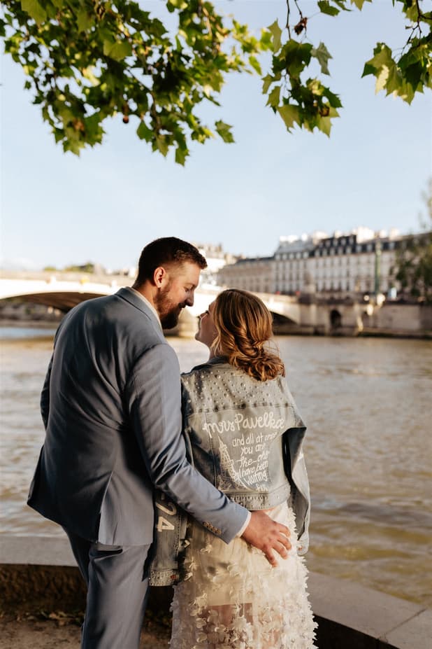 Bride and groom stand by the Seine under tree leaves, with a bridge and sunlit Paris buildings behind