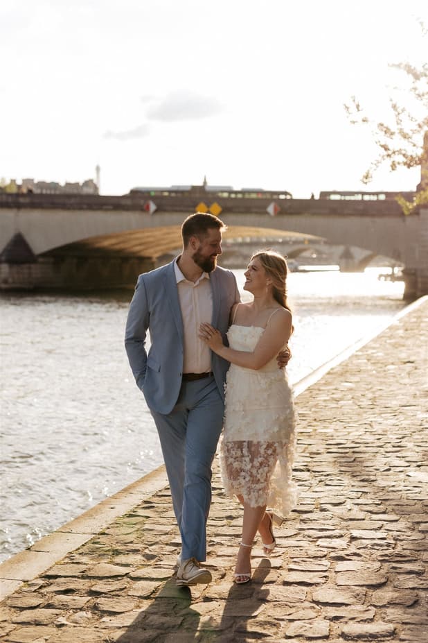 A bride and groom walk arm in arm along a riverside cobblestone path at sunset beside a bridge