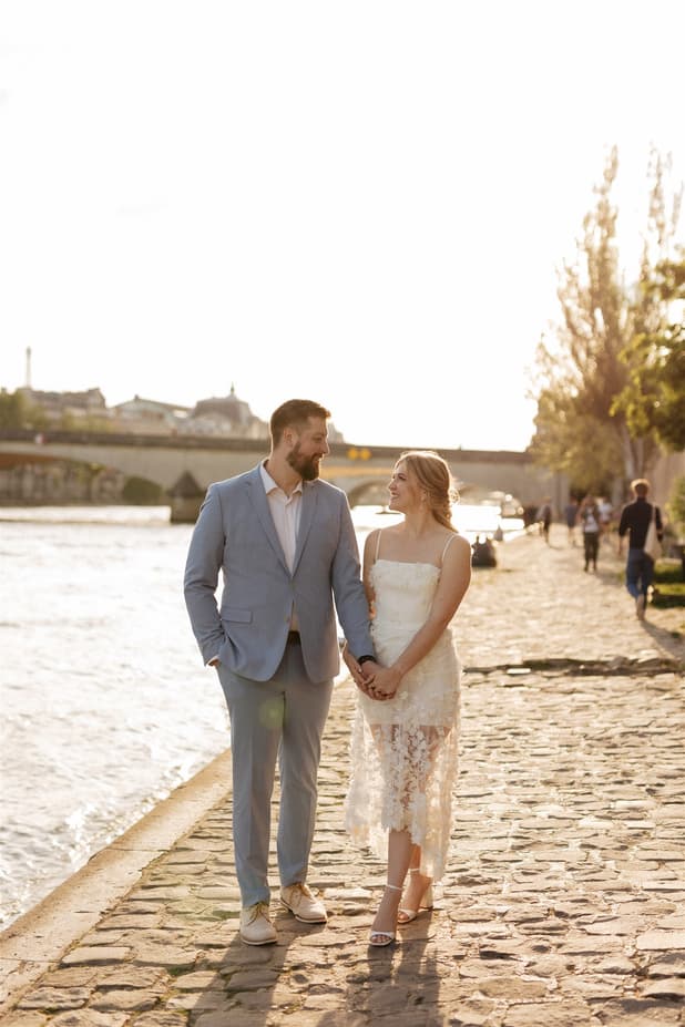 Bride and groom hold hands on a sunny Seine riverbank beside a stone bridge and golden trees